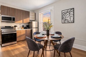 A modern kitchen with built-in appliances, a dining area with a round table and chairs, and a window with a view of the sunset. - The Penn Hotel - Steps to Gaslamp & Petco Park, AC (San Diego)