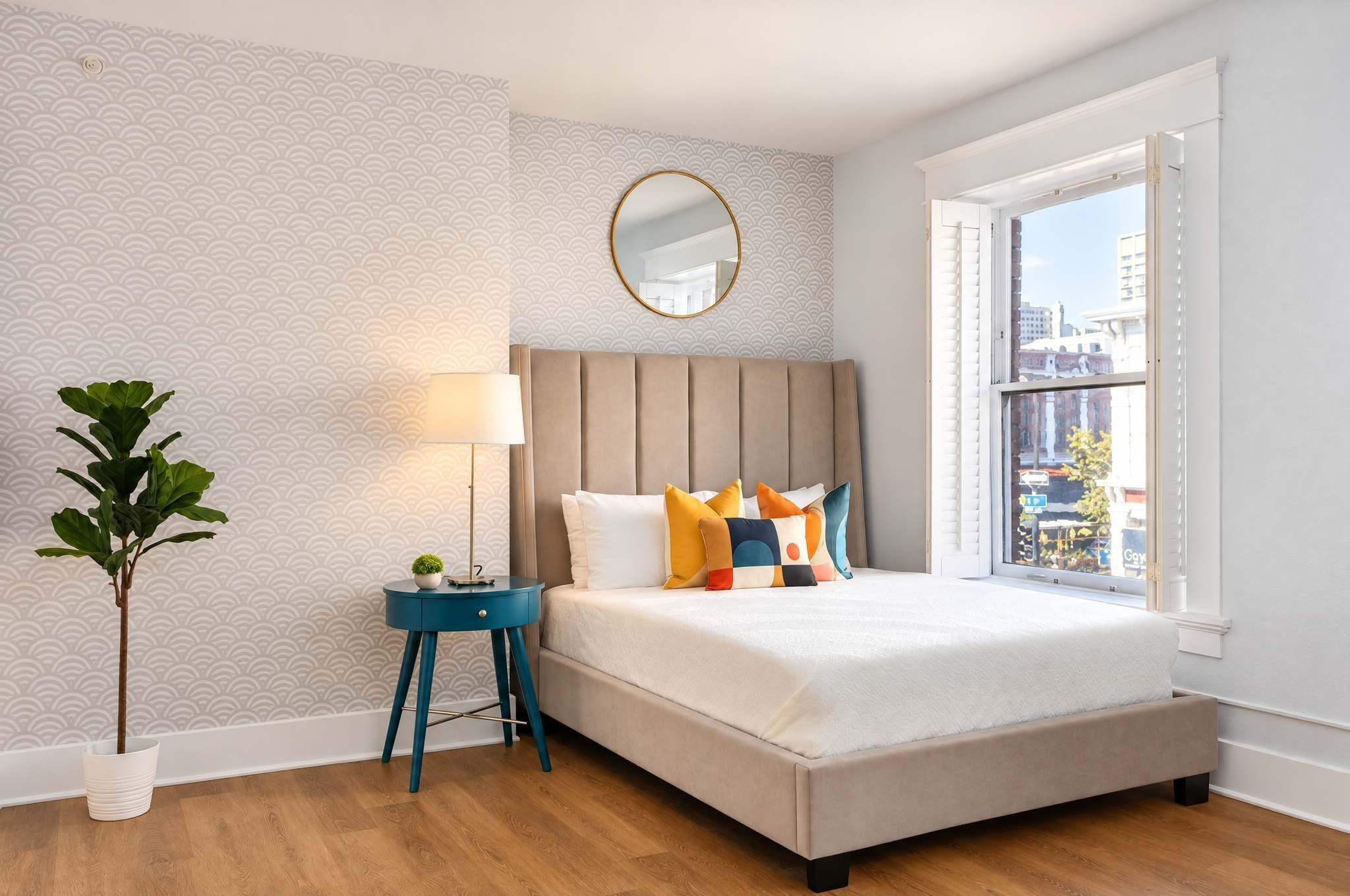 A bedroom with a bed, a bedside table with a lamp, a potted plant, and a window with a city view.