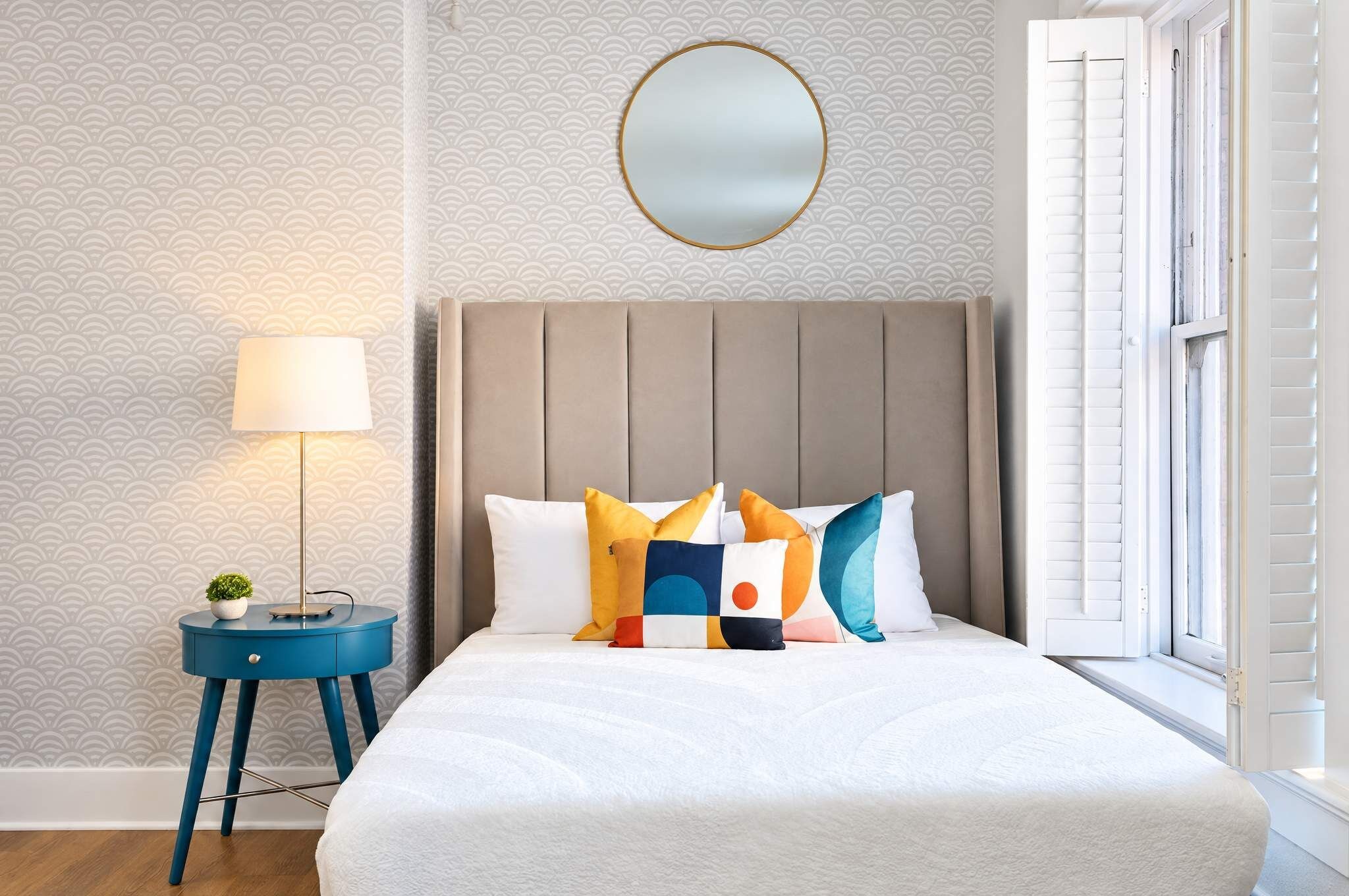 A bedroom with a bed, a bedside lamp, a round mirror, and a window with shutters.