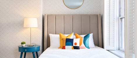 A bedroom with a bed, a bedside lamp, a round mirror, and a window with shutters.