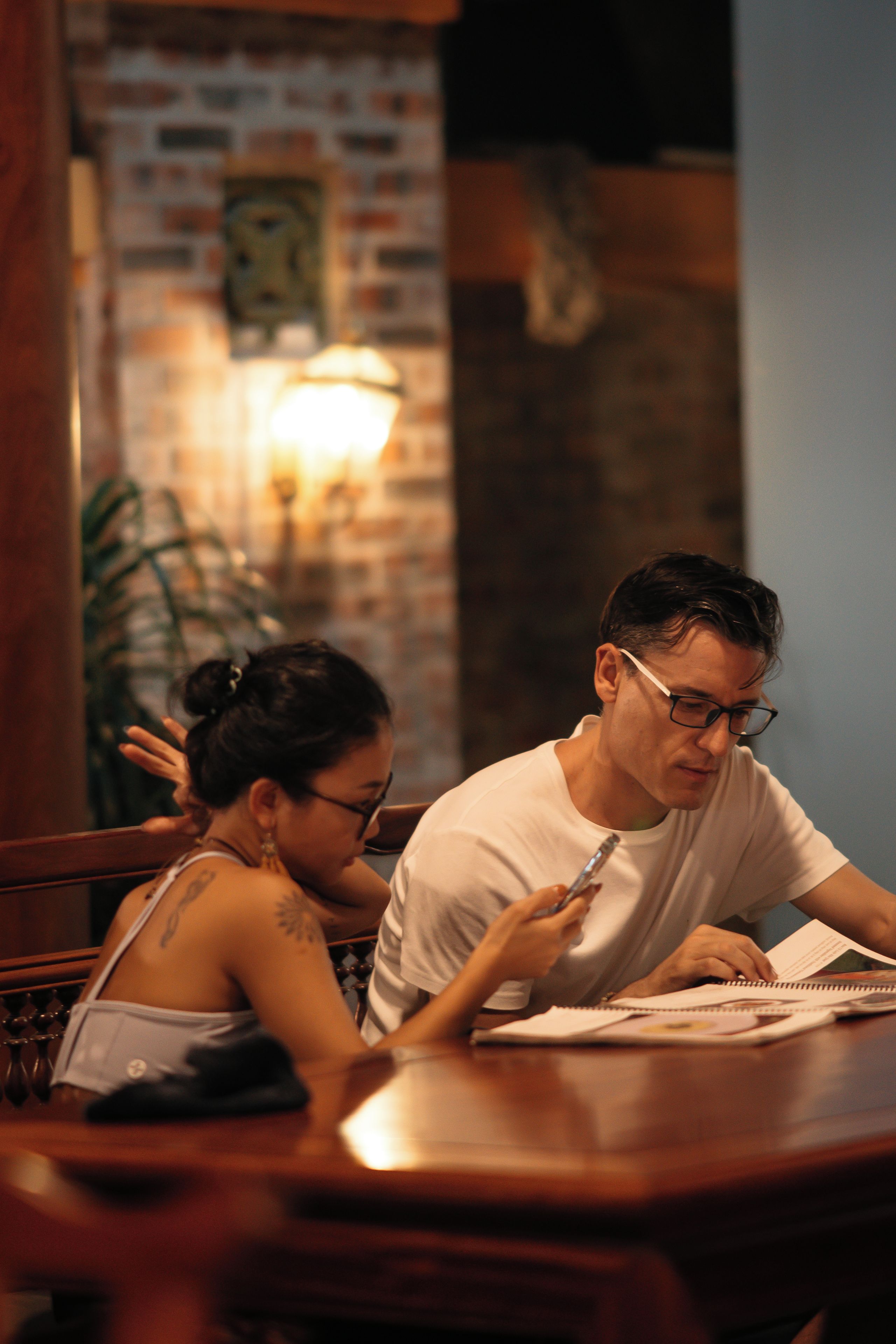 A person sitting at a wooden table with a notebook and pen, in a room with brick walls and a hanging light fixture.