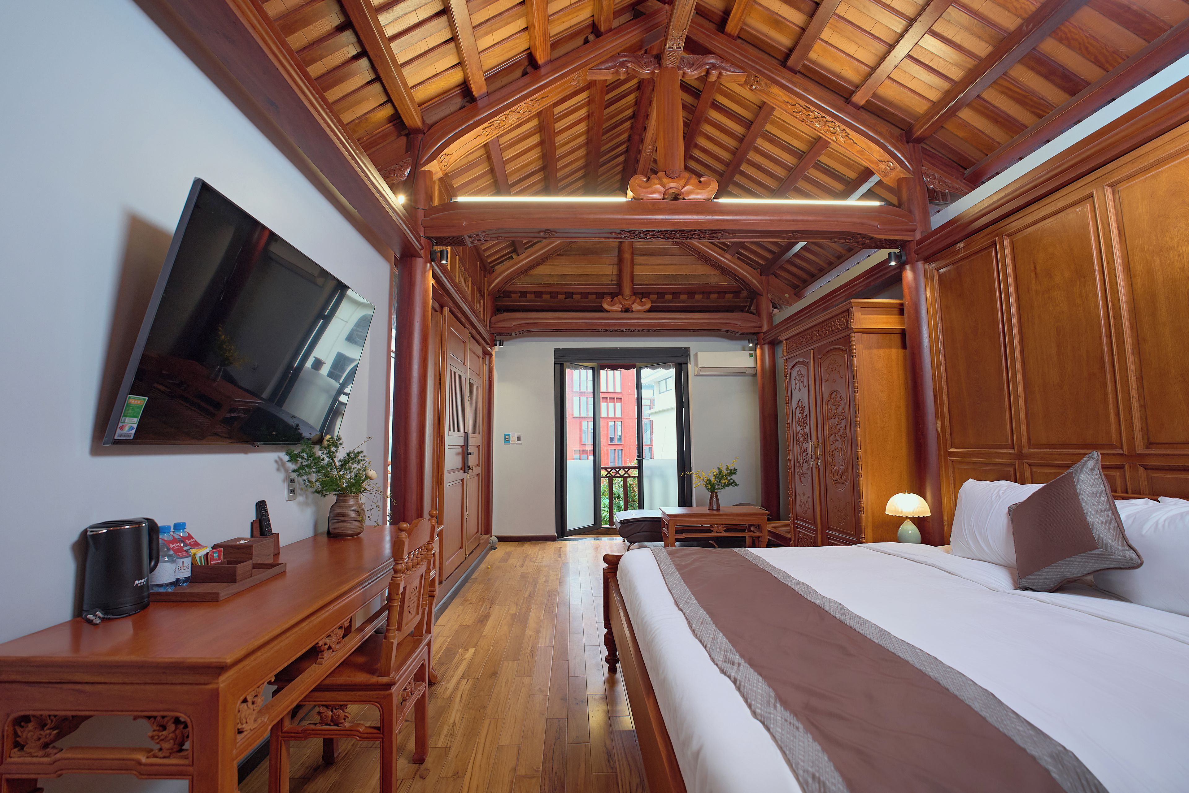 Une chambre d’hôtel avec un plafond en bois, un lit, un téléviseur fixé au mur et un bureau agrémenté d’un vase de fleurs.
