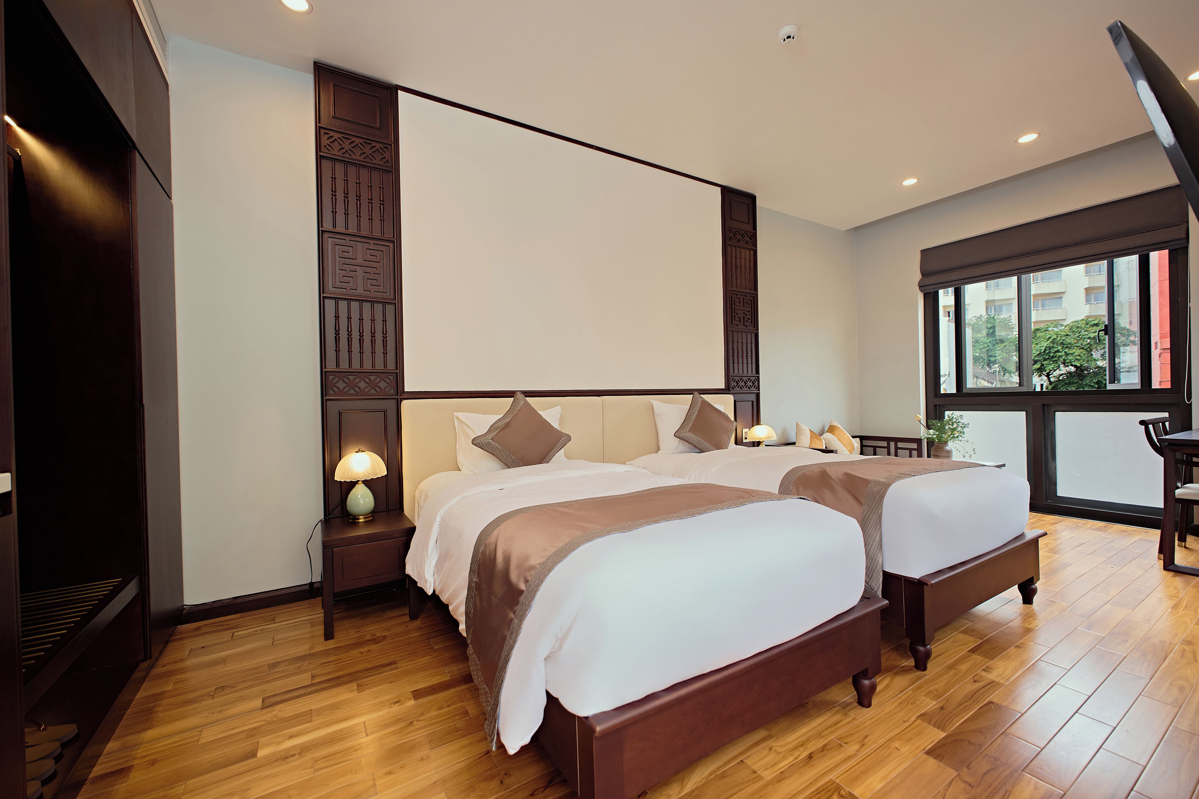 A hotel room with two beds, wooden flooring, a large window, and a view of the outside.