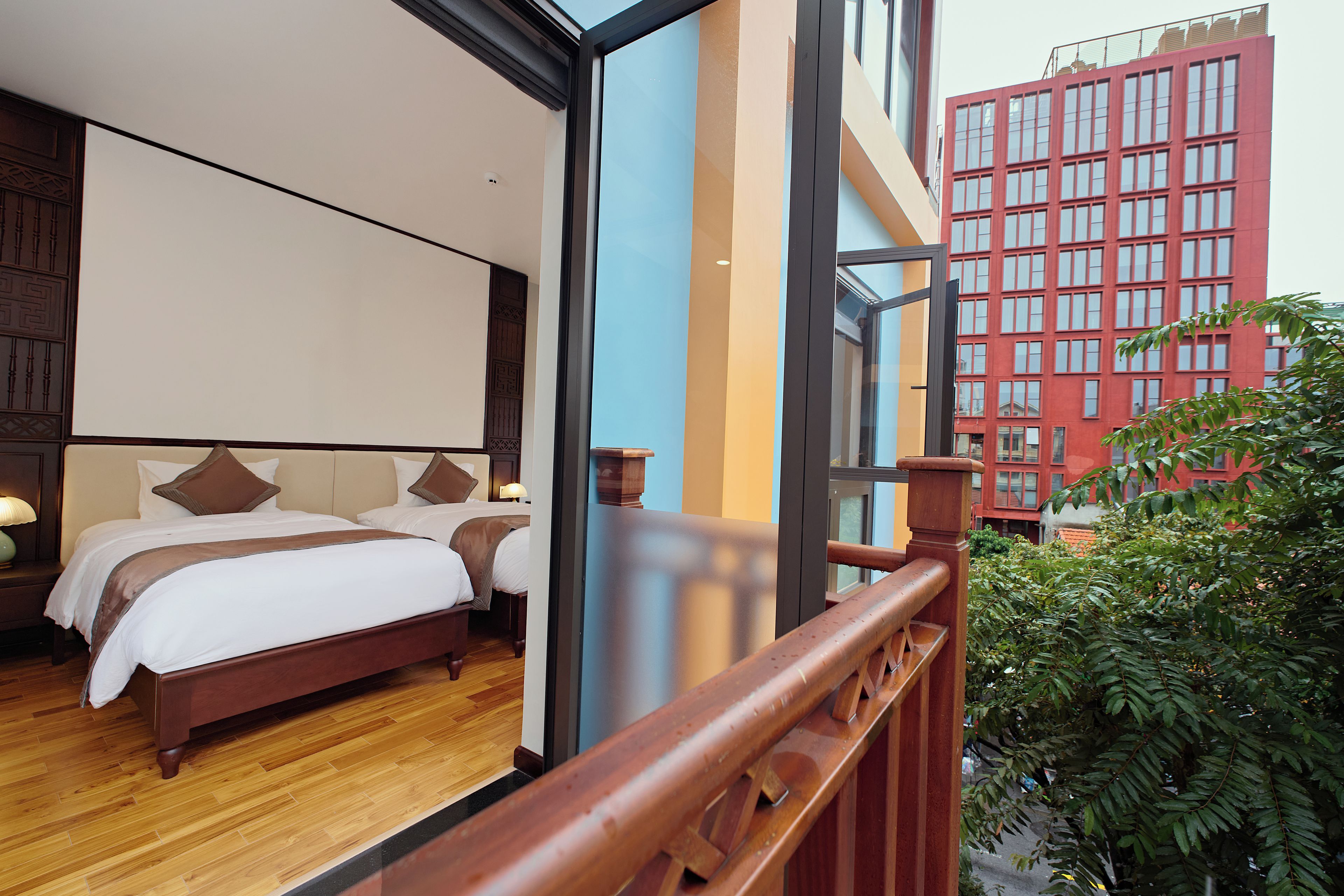 A hotel room with a large bed, wooden floor, and a view of a red building through a glass door.