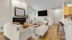 Una sala de estar moderna con chimenea, televisor de pantalla plana en la pared y una zona de cocina con armarios de madera y taburetes de bar.