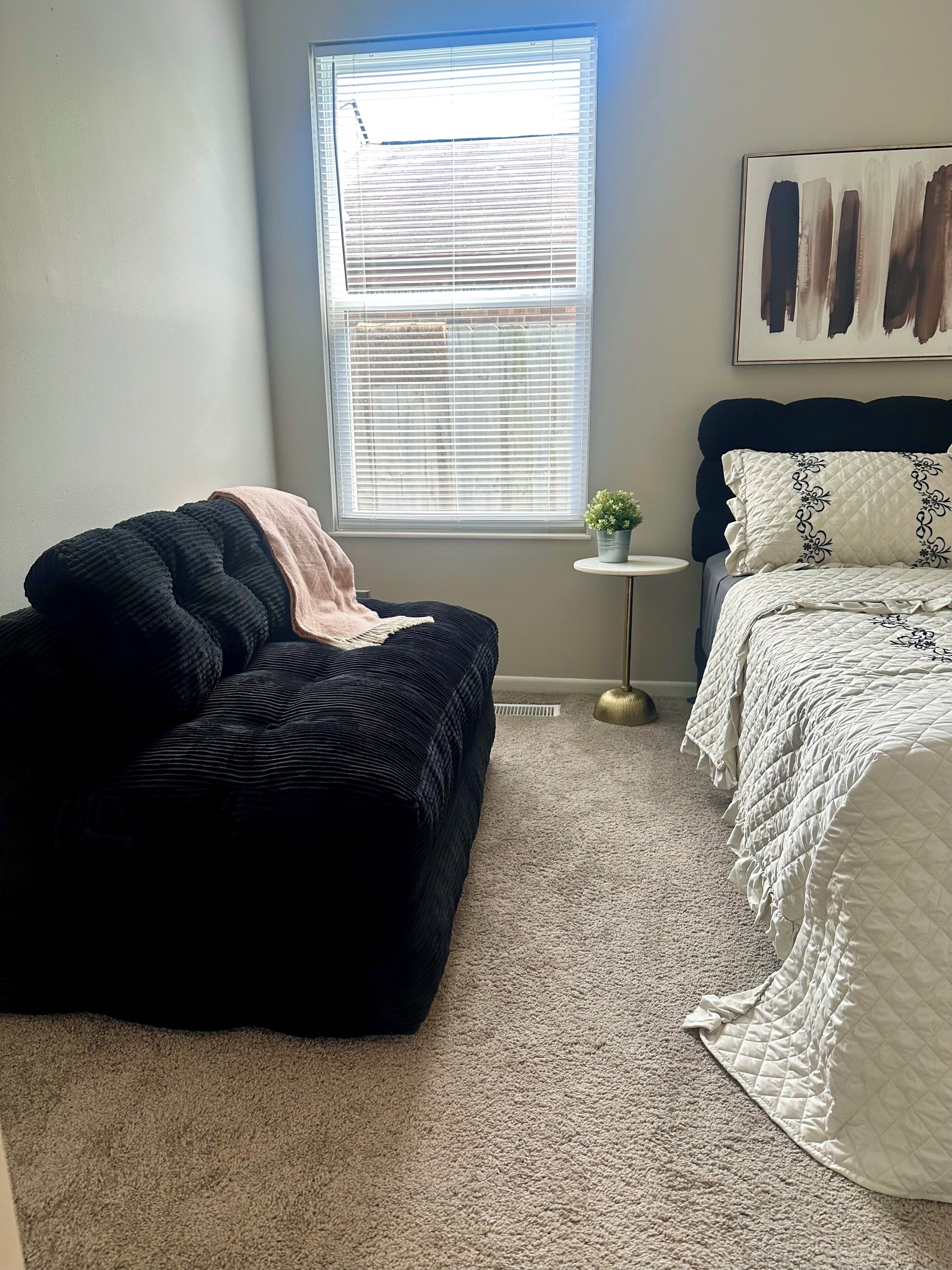 A bedroom with a bed, a window with blinds, a small table with a plant, and a black ottoman with a blanket.
