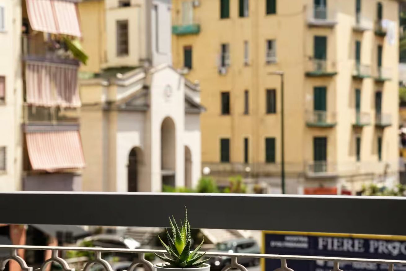 Un balcón con una maceta, con vista a una calle con edificios y un letrero.
