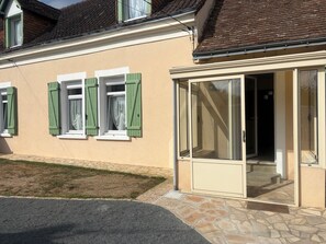 Ein Haus mit hellbeiger Fassade, grünen Fensterläden und einer Glastür, die zu einer überdachten Veranda führt.