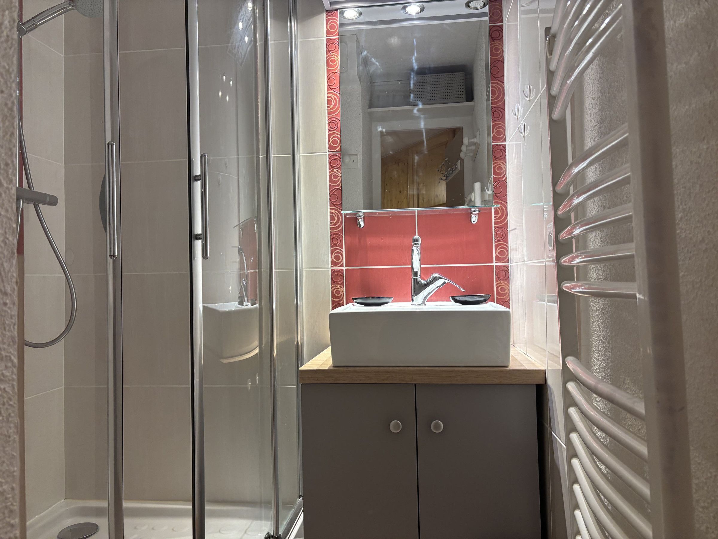 A modern bathroom with a glass-enclosed shower, a white sink with a red-tiled backsplash, and a wooden vanity.