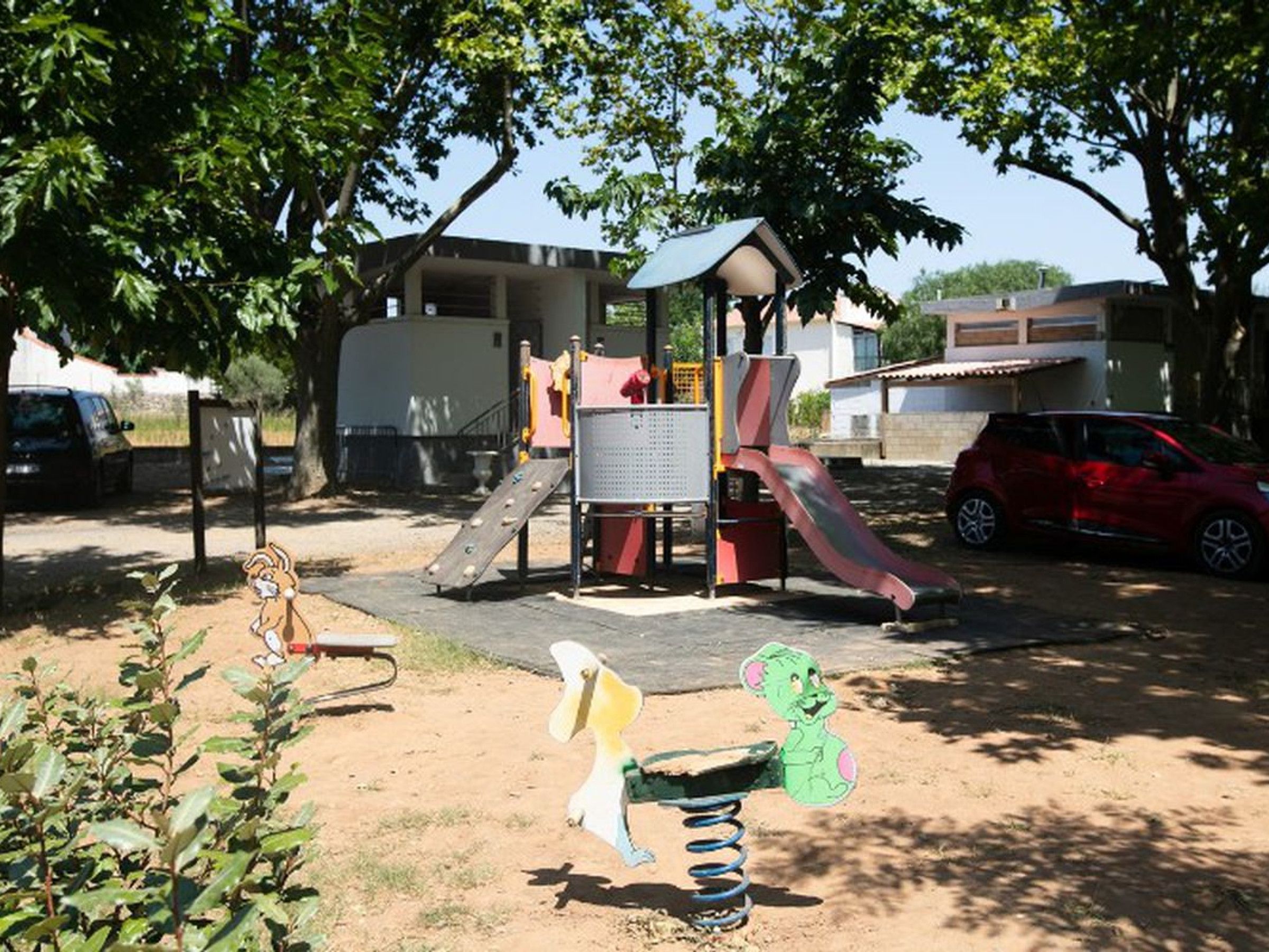 Parquinho com escorregador e estruturas de escalada, cercado por árvores e carros estacionados.