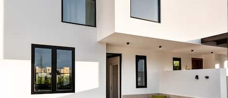 Una casa moderna de dos pisos con techo plano, ventanas amplias y un pequeño patio con mobiliario exterior.