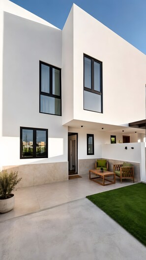 Une maison moderne à deux étages, dotée d’un toit plat, de grandes fenêtres et d’un petit patio aménagé avec du mobilier d’extérieur.