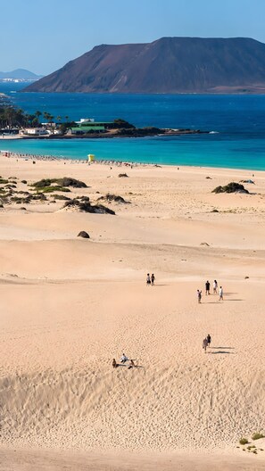 Ein Strand mit Menschen, die spazieren gehen und sitzen, ein klarer Himmel und ein Berg im Hintergrund.