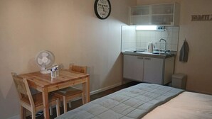 A small, compact room with a bed, a wooden table with a fan, a kitchenette, and a wall clock. - Comfortable Family Room with Breakfast at the Farm (Grandrupt-de-Bains)