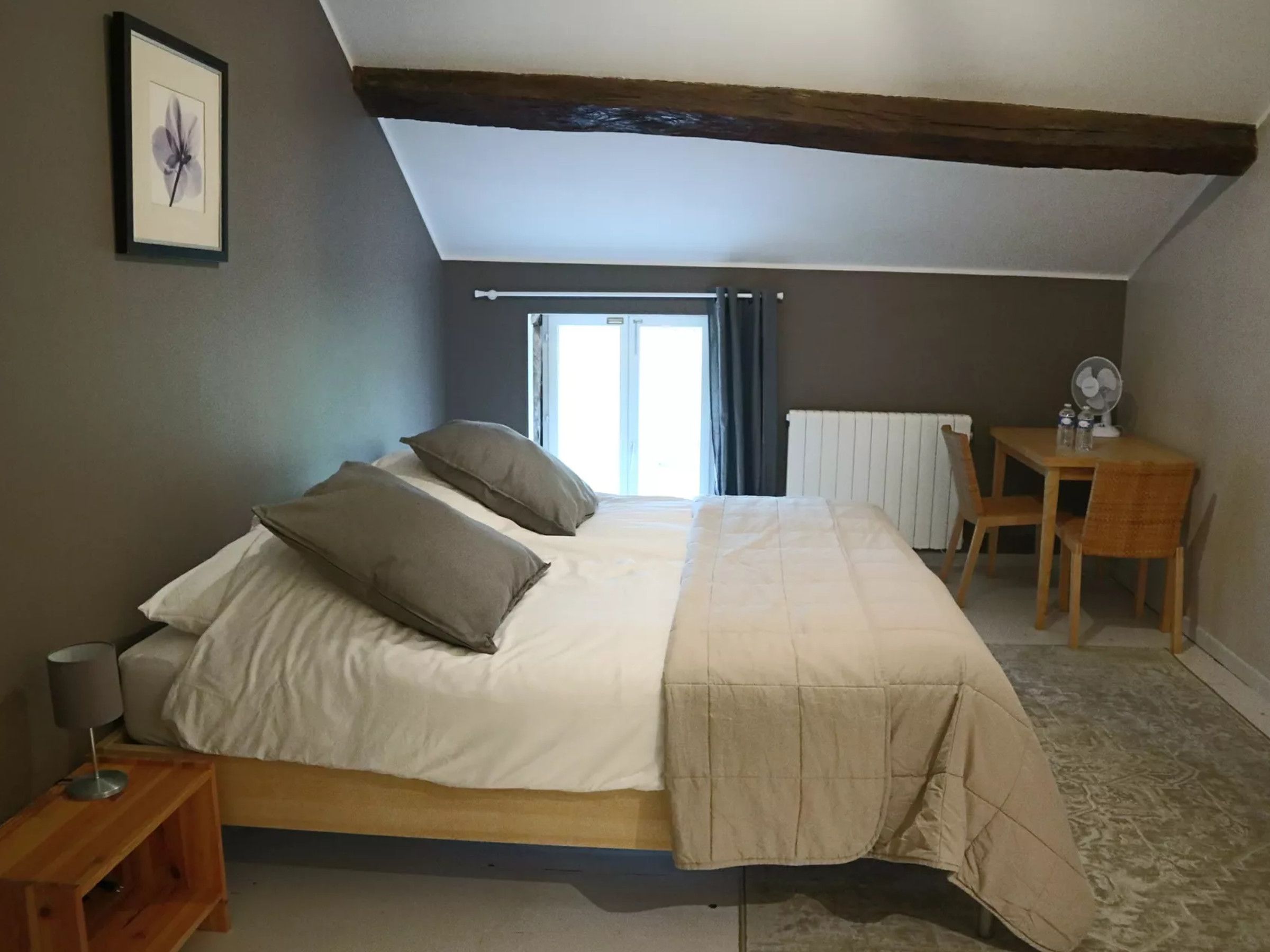 Et soveværelse med seng, natbord, stol, skrivebord med ventilator og et vindue med gardiner.