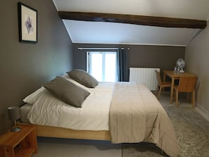 Un dormitorio con cama, mesita de noche, una silla, escritorio con ventilador y ventana con cortinas.