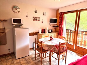A compact kitchen with a refrigerator, microwave, and a dining table with chairs. There is a window with a view of greenery outside. - Cozy apartment at the foot of the slopes - 2 guests (Saint-Jean-d'Aulps)