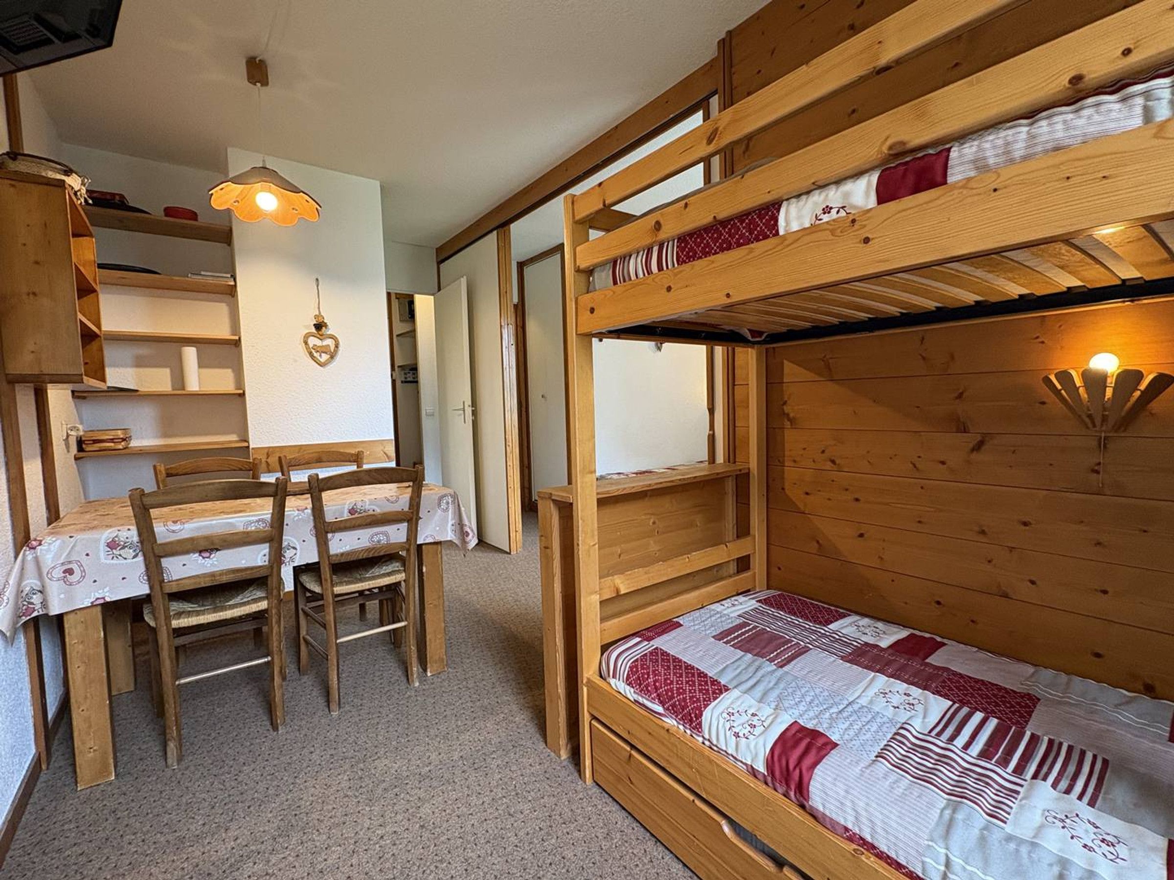 A bunk bed with a quilt, a wooden table with chairs, and a hanging light fixture.
