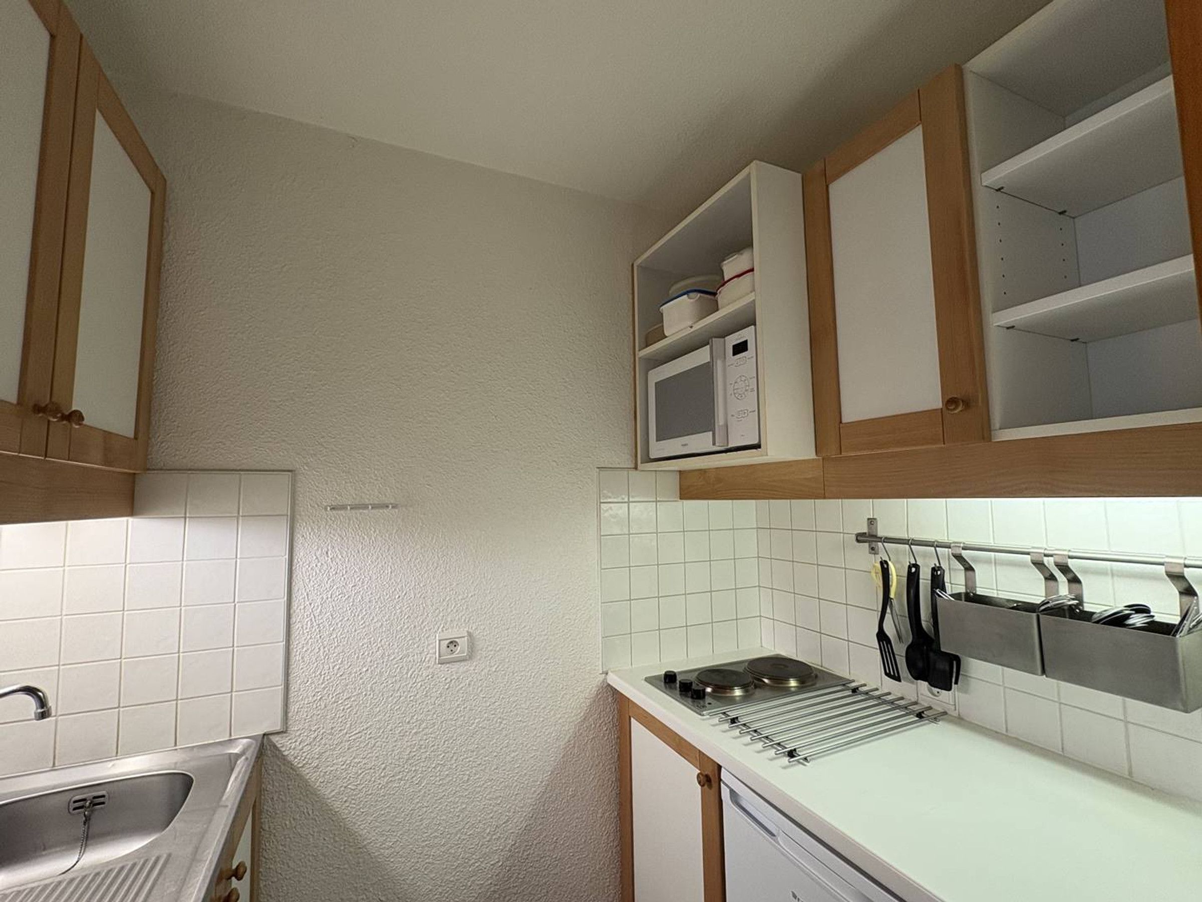 A compact kitchen with white tiles, a stainless steel sink, and wooden cabinets.
