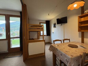 A room with a bunk bed, a dining table with chairs, a television mounted on the wall, and a view of the outdoors through a window.