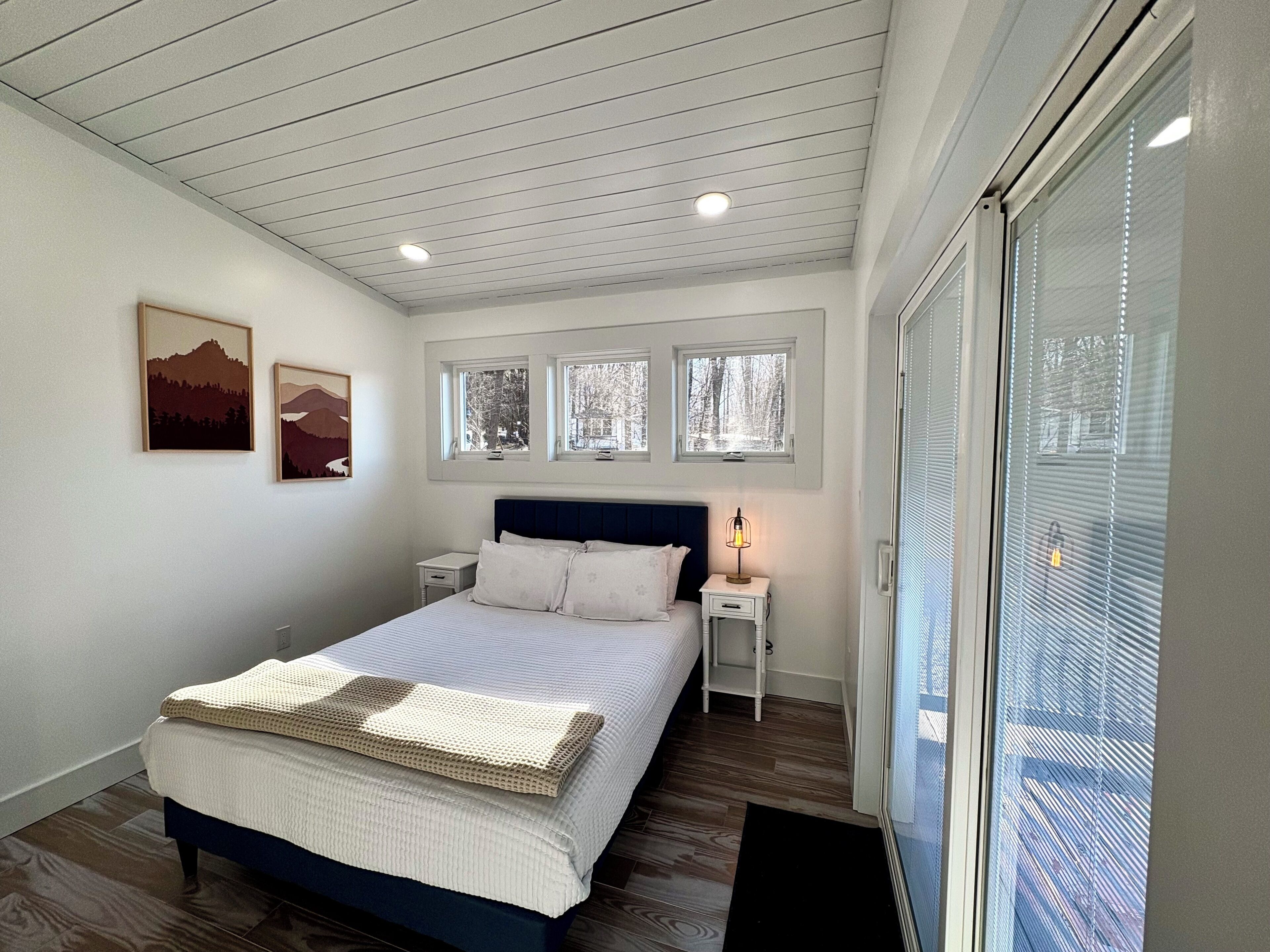 Un dormitorio con una cama, mesitas de noche, una lámpara y ventanas.
