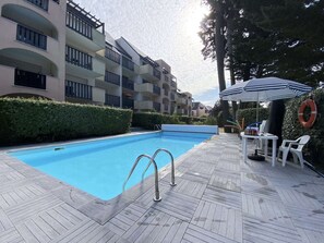 Una piscina con bordo in legno, circondata da un siepe e un edificio a più piani con balconi.