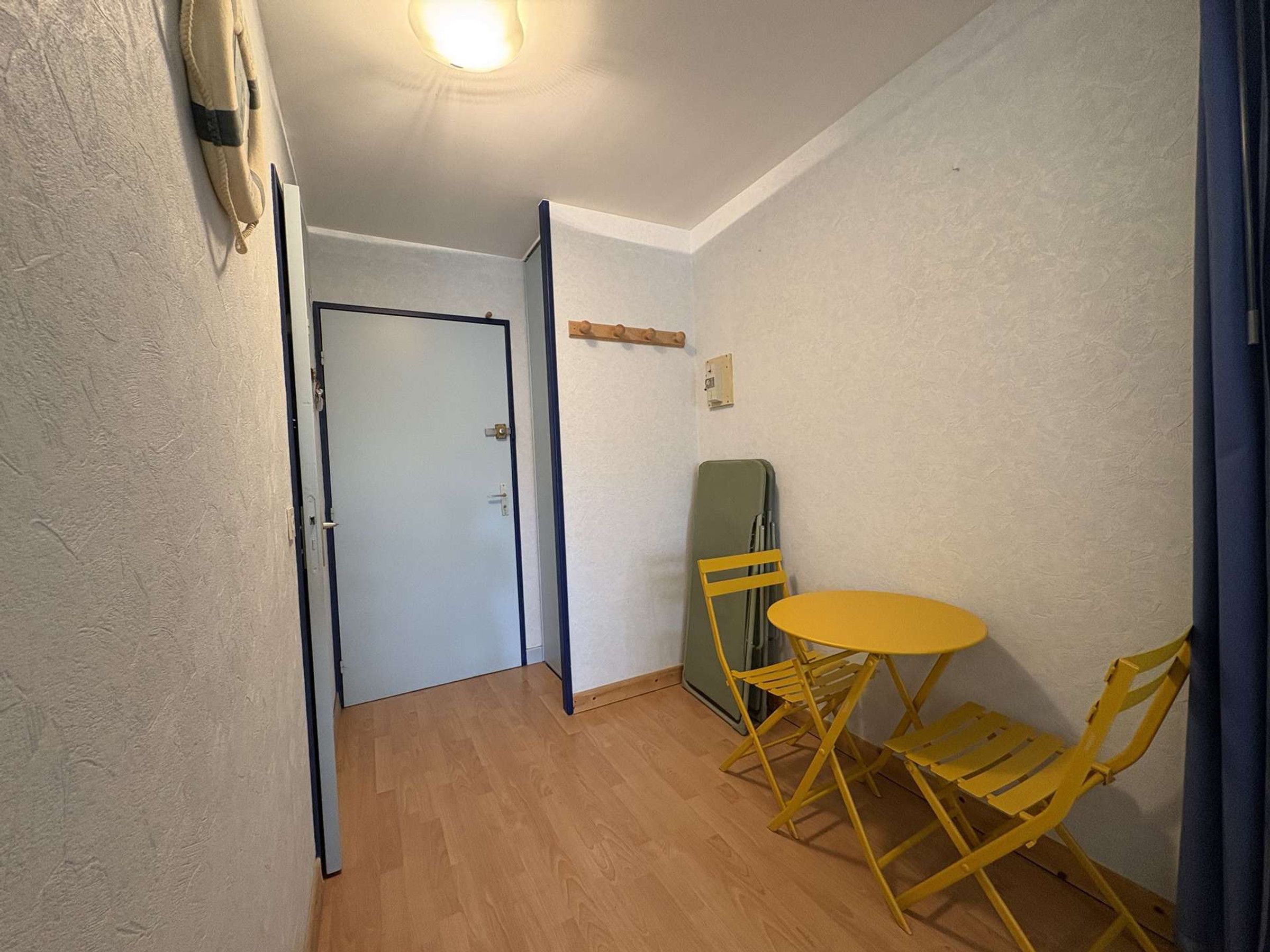 Une petite pièce avec un plancher en bois, une table pliante jaune et deux chaises jaunes. On aperçoit une porte au fond.