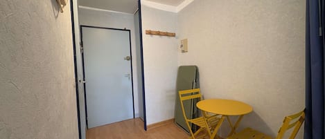 Una habitación pequeña con piso de madera, una mesa plegable amarilla y dos sillas amarillas. Se ve una puerta al fondo.