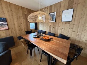 Un comedor con una mesa de madera, sillas negras, una lámpara colgante tejida y un bol de naranjas.