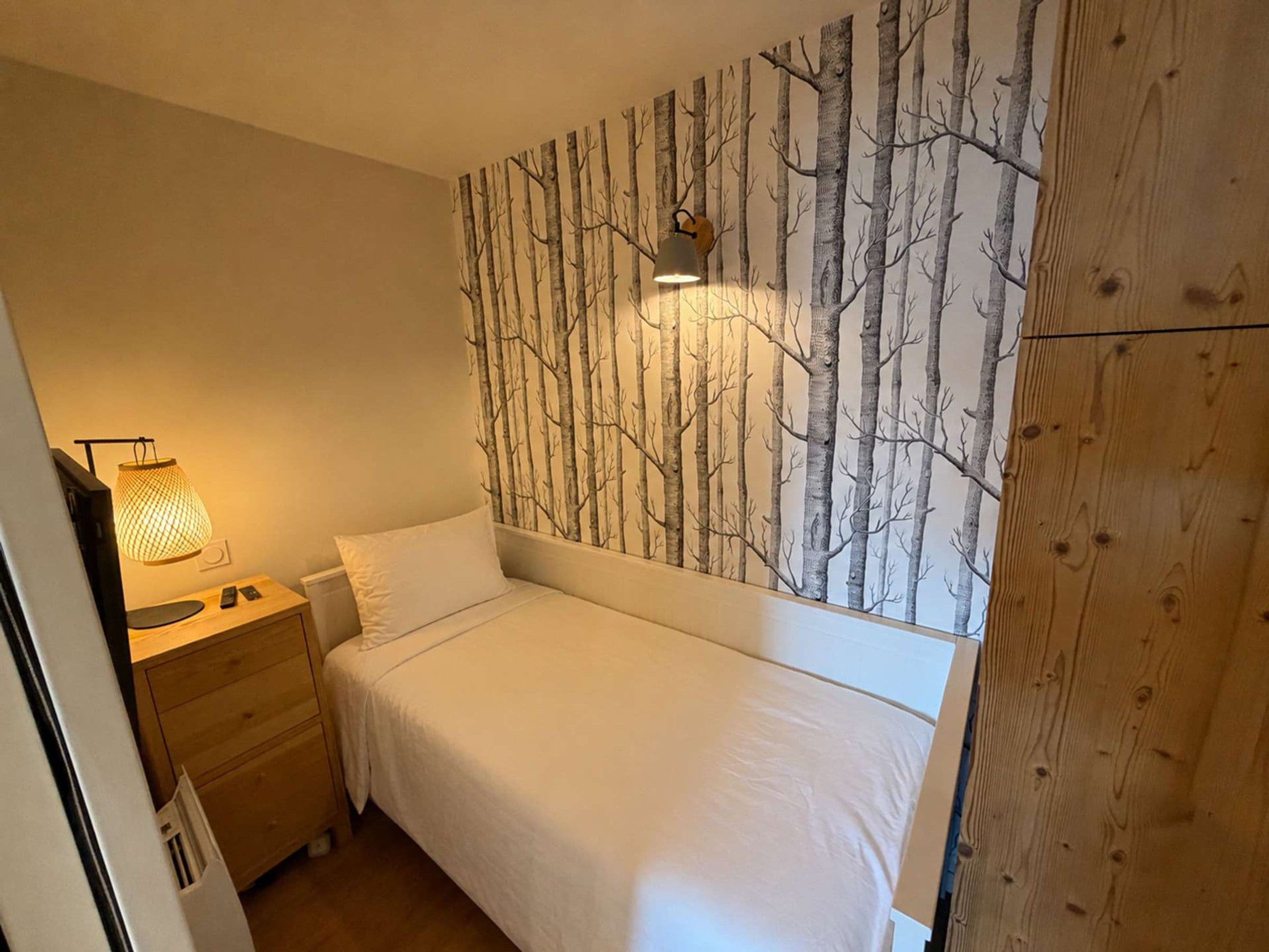 Un dormitorio con cama, mesita de noche, lámpara y un papel tapiz con diseño de árbol.