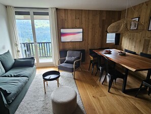 Ein modernes Wohnzimmer mit einer Couch, einem Esstisch und einem Sessel. Blick auf einen Balkon mit Geländer.