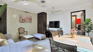 Un salon moderne comprenant un canapé, un pouf, une table à manger, des chaises, un téléviseur, des ventilateurs de plafond et un élément décoratif mural.