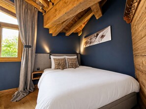 A bedroom with a bed, wooden ceiling beams, a nightstand, a window with curtains, and a wall-mounted light fixture. - Comfortable apartment valley view - 10 guests (Saint-Martin-de-Belleville)