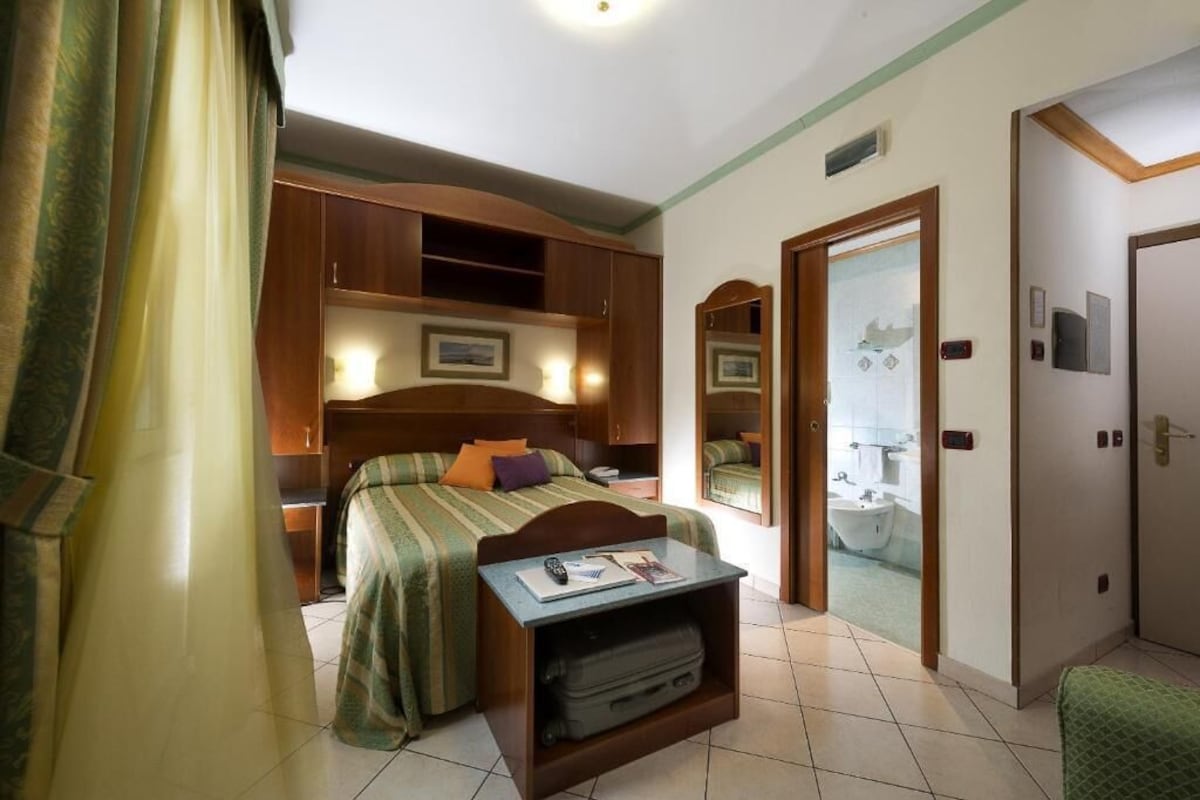 Une chambre d’hôtel avec un lit, un bureau sur lequel se trouve une valise, un miroir et une salle de bain visible à travers une porte ouverte.