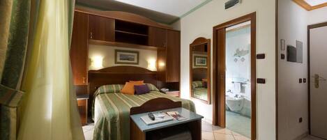 A hotel room with a bed, a desk with a suitcase, a mirror, and a bathroom visible through an open door.