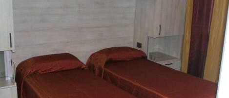 A room with two beds, each with a red bedspread, a white nightstand, and a small table.