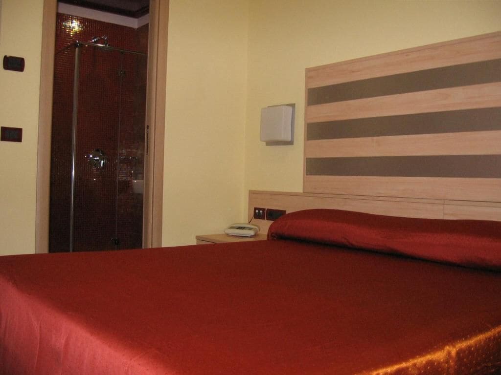 Una habitación de hotel con cama, cabecera de madera, mesita de noche, teléfono y zona de ducha.