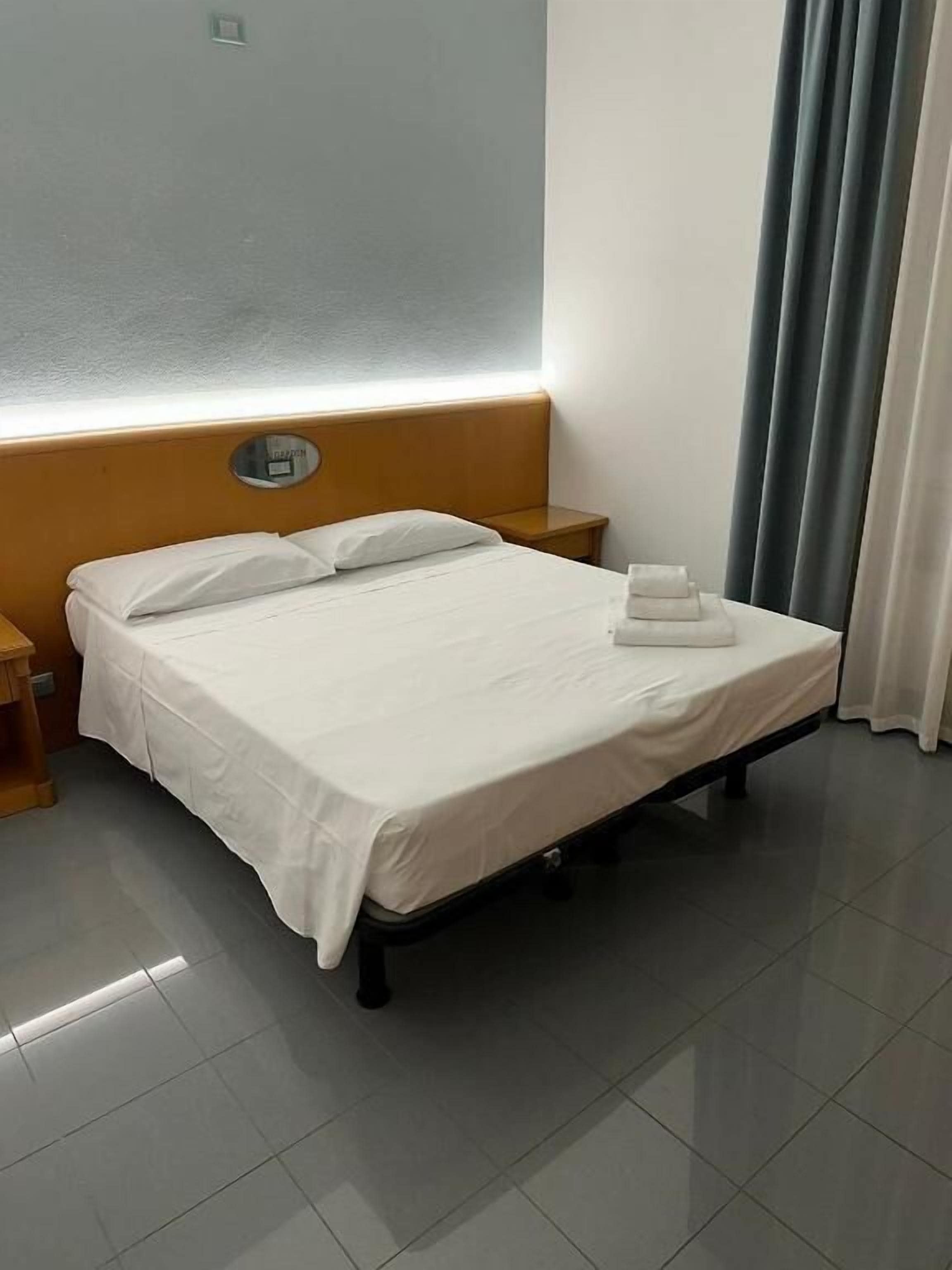 Hotellihuone, jossa on sänky, yöpöytä, seinälle kiinnitetty kello ja verho.