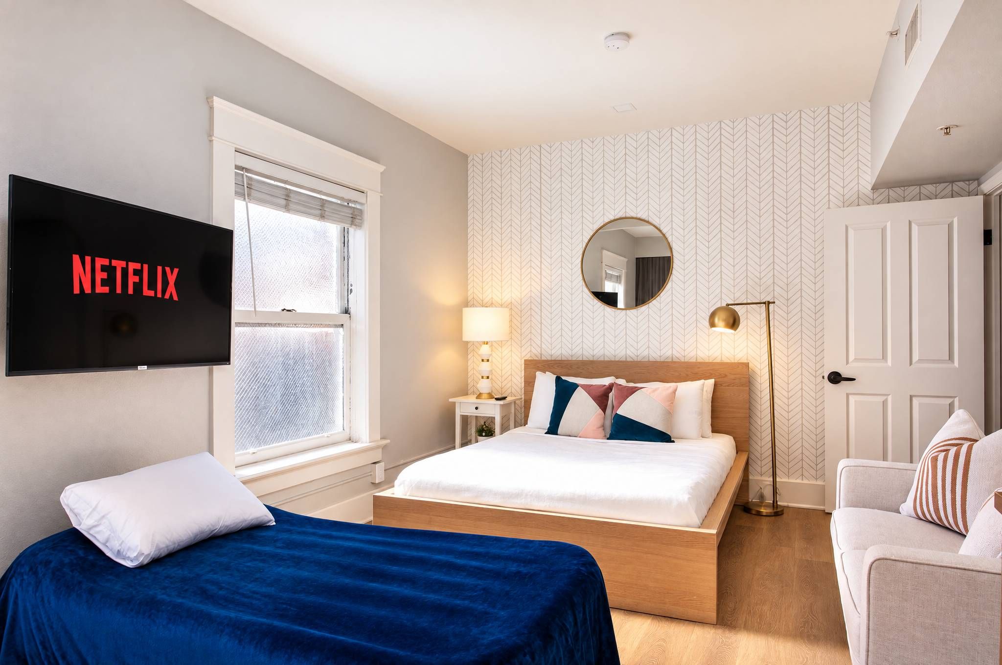 Habitación de hotel con cama, televisor, mesita de noche, lámpara, espejo y una silla.