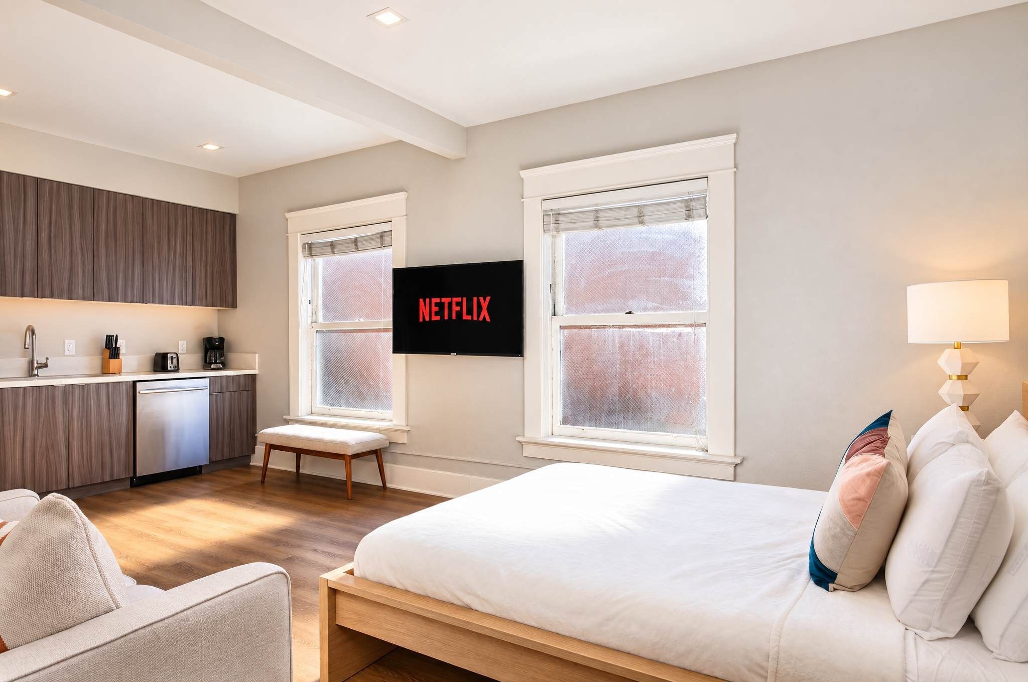 A modern hotel room with a bed, a seating area, a kitchenette, and a TV mounted on the wall.