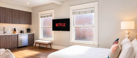 A modern hotel room with a bed, a seating area, a kitchenette, and a TV mounted on the wall.