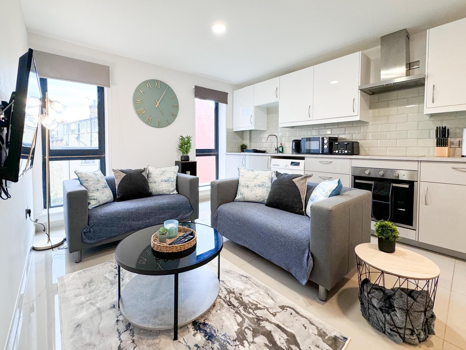A modern living room with two sofas, a coffee table, and a kitchen area with white cabinets and appliances.