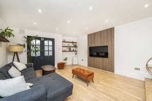 A modern living room with a grey sofa, a brown ottoman, a TV mounted on a wooden panel wall, and a door with frosted glass panels. - Luxury 2 bed maisonette in Spitalfields zone 1 - Pass the Keys (London)