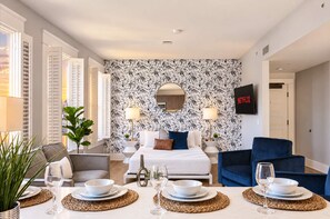 A modern bedroom with a large bed, a TV, and a dining area with a table set for two. - The Penn Hotel - Designer 2BR Near Gaslamp & Park (San Diego)