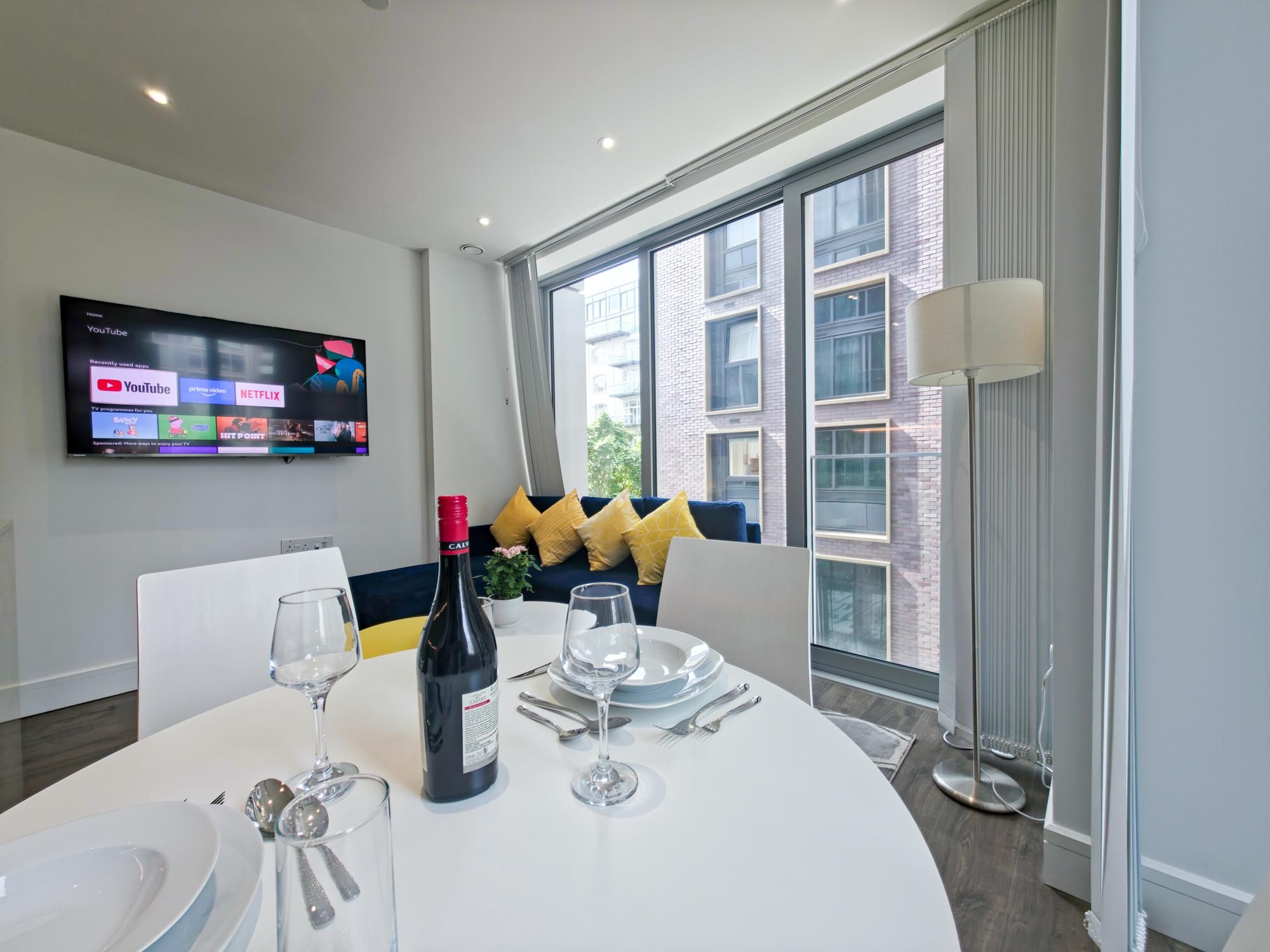 Een moderne eetruimte met een ronde tafel gedekt voor twee personen, een aan de muur bevestigde televisie die streamingdiensten toont, en een groot raam met uitzicht op de stad.