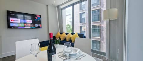 Un comedor moderno con una mesa redonda para dos personas, un televisor en la pared mostrando servicios de transmisión, y un ventanal con vistas a la ciudad.