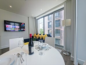 A modern dining area with a round table set for two, a television mounted on the wall displaying streaming services, and a large window offering a view of the city. - Sleek flat near the City 5 min from station - Pass the Keys (London)