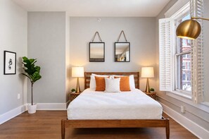 A neatly arranged bedroom with a wooden bed, two bedside lamps, a large window with shutters, and a framed artwork on the wall. - The Penn Hotel - King Bed, AC & Gaslamp Nightlife! (San Diego)