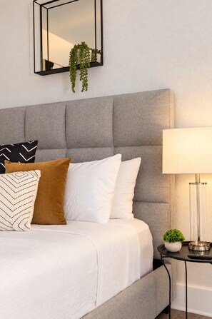A bedroom with a grey upholstered headboard, a bed with white and brown pillows, a bedside table with a lamp, and a hanging plant. - The Penn Hotel - Smart TV & King Bed Near Gaslamp! (San Diego)