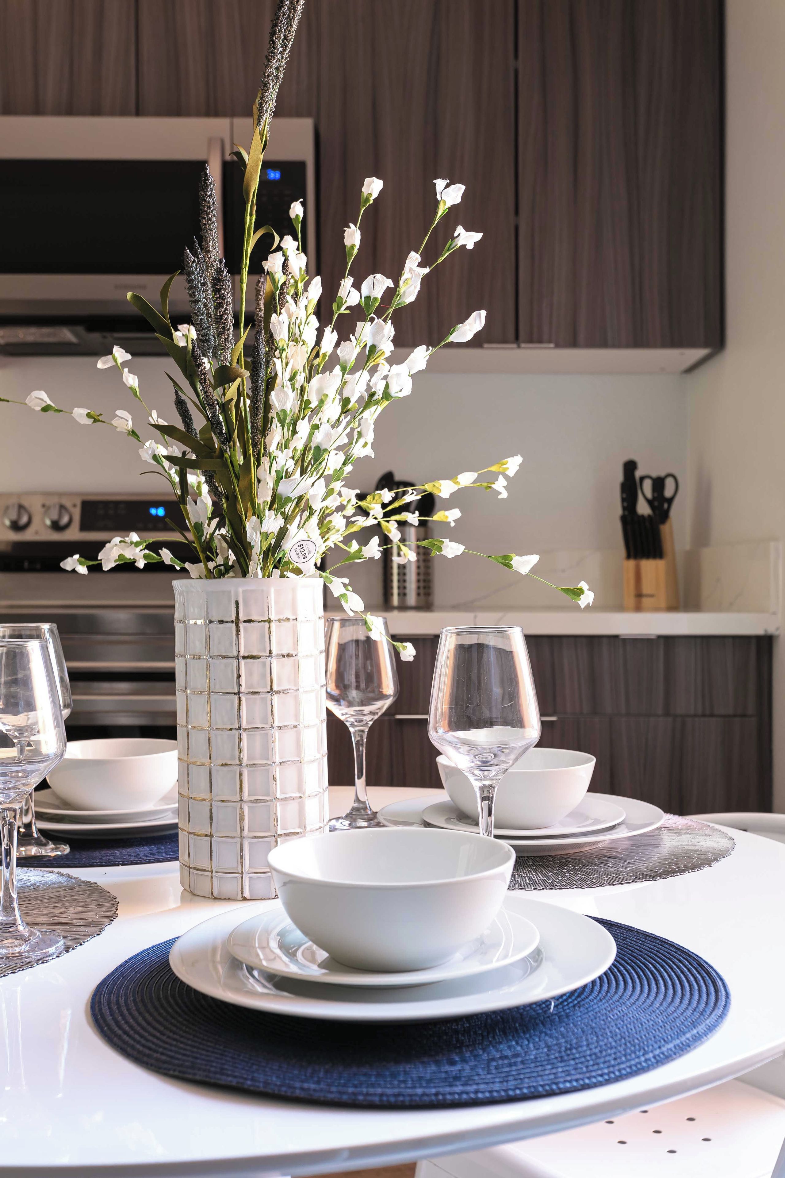 A modern dining table set with white bowls and wine glasses, a decorative vase with white flowers, and a kitchen in the background.